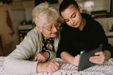 A teenage caucasian granddaughter shows her senior grandmother how to use a tablet. Education about...