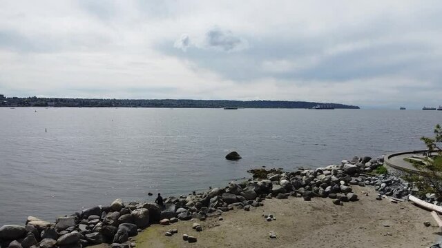 Aerial fly over rocky low tide English Bay Harbor next to the sea wall on an overcast spring day whereby theres no people nobody out for a walk and only one person suntanning on the rocks Vancity BC