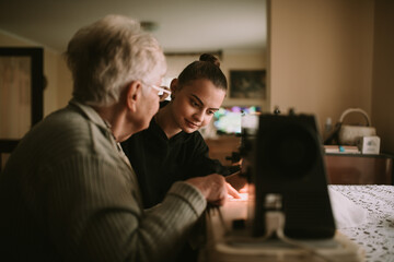 Senior sweet grandmother explains to her beautiful teenage granddaughter to sew clothes on a sewing machine in her grandmother's house