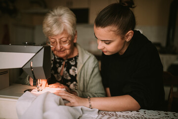 Smiling caucasian grandmother and her teenage granddaughter sew clothes together on a sewing machine in the living room