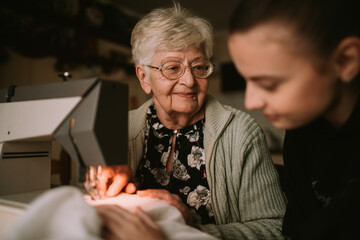 Senior cute smiling grandmother looks at her granddaughter as she teaches her to sew on a sewing machine in the living room