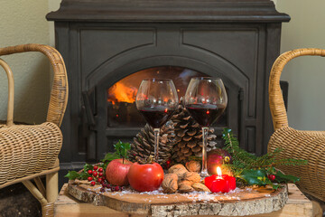Winter holidays still life. Two glasses of red wine, fruits, nuts, cones, fir branches on the plate in front of the burning stove