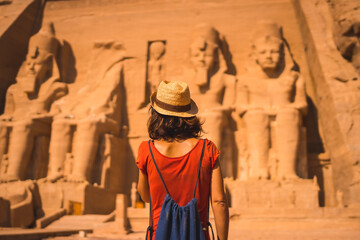 A young tourist in a red dress and straw hat walking towards the Abu Simbel Temple in southern Egypt in Nubia next to Lake Nasser. Temple of Pharaoh Ramses II, travel lifestyle - obrazy, fototapety, plakaty
