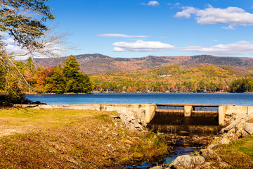 Colorful fall foliage and the Keewaydin Dam along the shore of Keewaydin Lake in Stoneham, Maine with mountains in the background above a bright blue sky.
