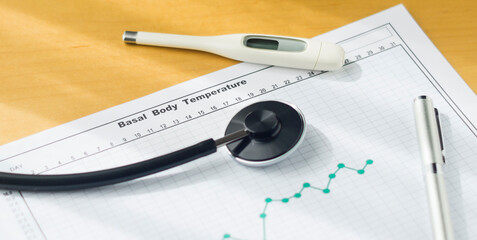 A thermometer, a pen, a stethoscope and a Basal Body Temperature Chart  on the desk.