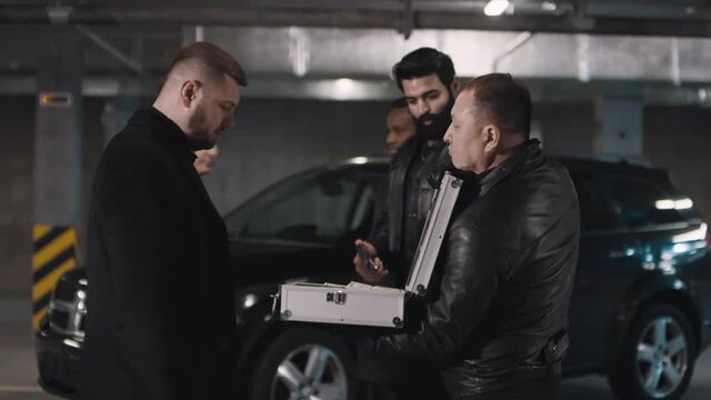 Medium shot of bandits standing in row opening metal briefcase showing money to other criminals standing at isolated underground parking