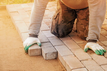 man in gloves lays paving stones in layers on garden pathway.  Laying gray concrete paving slabs in courtyard on leveled sand foundation base. Road Paving, construction, repairing sidewalk.