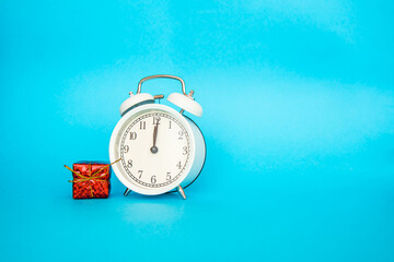 White alarm clock on a blue background. Free space for an inscription. Time concept. White alarm clock with small red new year gift box