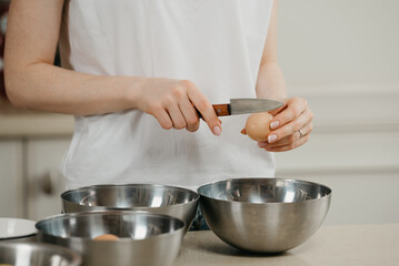 A photo of the hands of a young woman who is breaking the eggshell of the organic farm egg with the knife above the stainless steel soup bowl in the kitchen