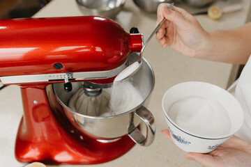 A close photo of the hands of a young woman who is spooning sugar to stainless steel soup bowl with the egg whites while the red stand mixer is whipping it.
