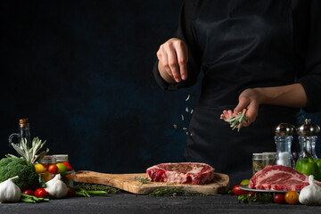 Professional chef in black uniform pours rosemary on raw steak on wooden chopped board. Backstage of preparing grilled pork meat at restaurant kitchen on dark blue background. Frozen motion.