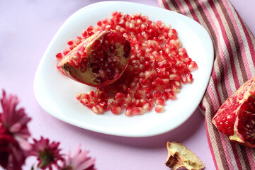 Pomegranate seeds with a cut piece of fruit lie on a white plate on a purple background, close-up, bokeh