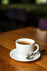 A white cup of black coffee, white saucer, spoon, wooden table, at a cafe