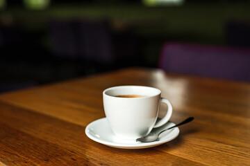 A white cup of black coffee, white saucer, spoon, wooden table, at a cafe