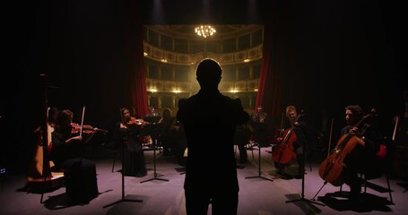 Cinematic shot of conductor directing symphony orchestra with performers playing violins, cello and trumpet on classic theatre with curtain stage during music concert with dramatic lights. - Powered by Adobe