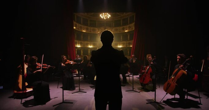 Cinematic shot of conductor directing symphony orchestra with performers playing violins, cello and trumpet on classic theatre with curtain stage during music concert with dramatic lights.