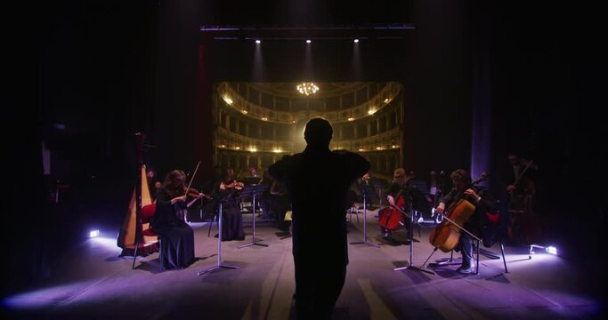 Cinematic shot of conductor directing symphony orchestra with performers playing violins, cello and trumpet on classic theatre with curtain stage during music concert with dramatic lights.