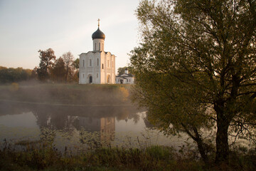 An XII century Russian Orthodox church above a misty pond lit by the rising sun. A tree in the foreground. Autumn colours. No people.