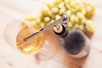 A glass of white wine and a bottle being opened are on a wooden table. A sommelier knife in the...
