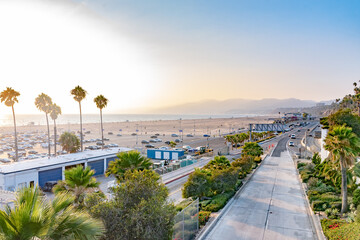 Scenic California coast view of beach, palm trees, mountains and highway. Miles of summer weather coastal city landscape. Vehicles drive in traffic, other cars parked on sand at Santa Monica Pier.