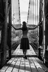 a young woman stands with her back to the old suspension bridge, black and white photo