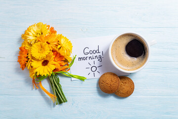 Notes good morning and coffee mug with bouquet of yellow flowers calendula on blue background. Breakfast, morning coffee, card concept, top view