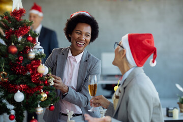 Cheerful black businesswoman and her colleague having fun while decorating Christmas tree in the...
