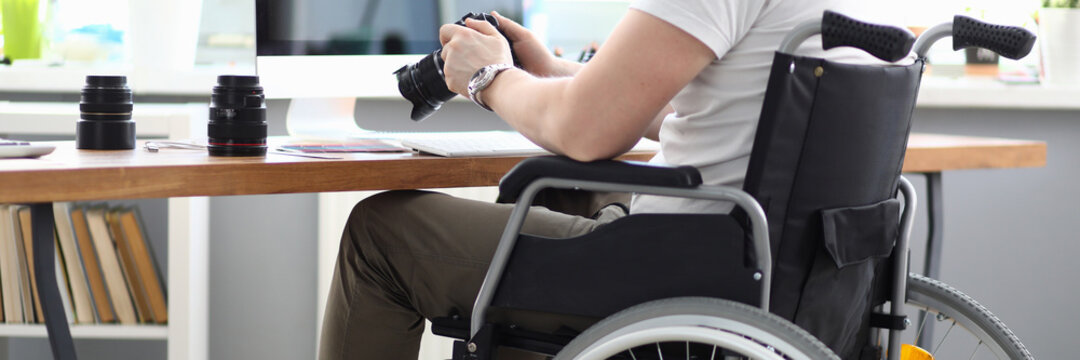 Close-up of professional man photographer holding camera and sitting in wheelchair. Modern office with laptop on desktop. Disabled people and photography business concept