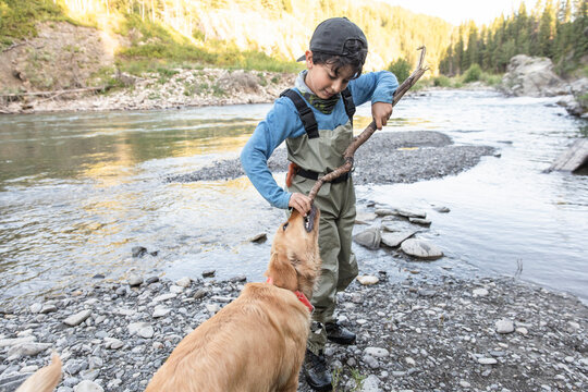 Boy wearing waders playing with pet dog by river