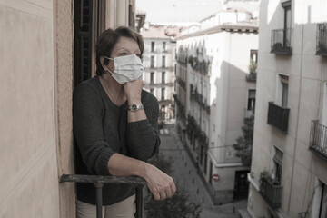 mature woman with face mask sad and scared at home balcony during covid19 pandemic lockdown looking to the street confused and depressed in senior people virus fear concept