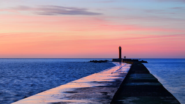 A couple of people standing near the orange lighthouse. Colorful sunset sky. Stunning cloudscape. Long exposure. National landmark, sightseeing, recreation theme. Baltic sea, Riga bay, Latvia