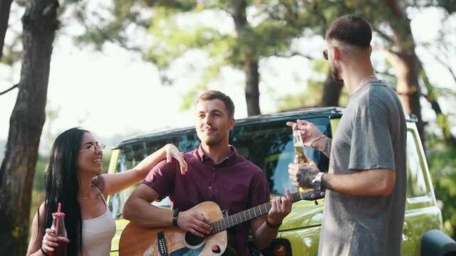 Three friends with American flan near modern green automobile plays guitar with drinks.
