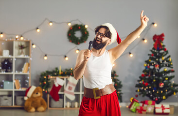 Funny smiling man Santa in festive home clothing celebrating Christmas and singing in microphone at...
