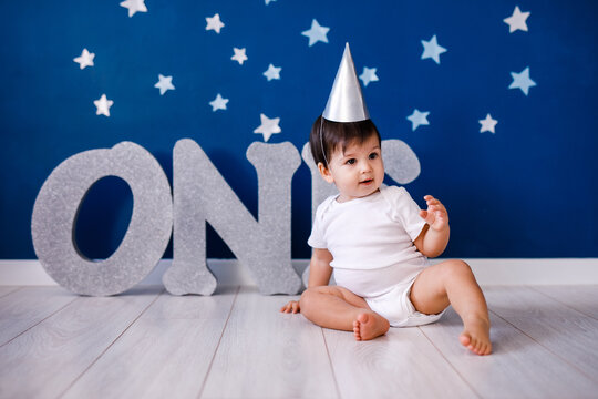 One year old baby boy in white t-shirt and silver hat celebrates birthday near silver letters ONE on purple background with stars