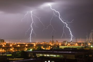 Powerful flash lightning hitting the city at night over dark gray sky. A strong lightning strike over hits the ground, illuminating the industrial area around.