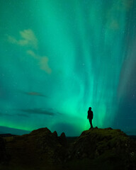 Naklejka na ściany i meble An adventure solo traveler posing with the beautiful northern lights also known as aurora borealis in the background. A breathtaking nature of Iceland as nordic country