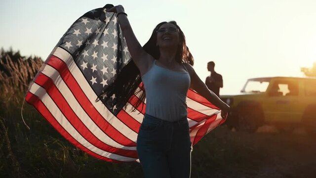 Group of friends standing with alcohol in hands near green automobile when girl dancing with American flag.
