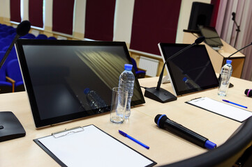 Conference room for business meeting, table with microphones, monitors, bottles with water, glasses, papers, pens and chairs, blue seats in row on blurred background, selective focus