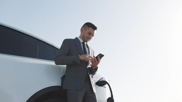 Caucasian man using smart phone and waiting power supply connect to electric vehicles for charging the battery in car. Plug charging an electrical car