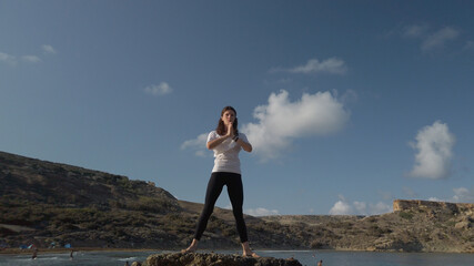 Obraz premium Young caucasian woman brunette doing yoga on rocky beach against blue sky. High quality photo