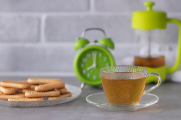 A bright green French press, a glass cup and saucer, cookies on a glass plate and a bright green alarm clock on a gray table.