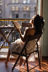 attractive woman covered in white sheet reading book and sitting on balcony