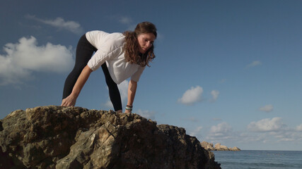 Obraz premium Young caucasian woman brunette doing yoga on rocky beach against blue sky. High quality photo