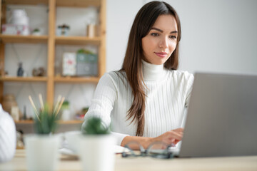Young woman in a white sweater works behind a laptop, blured foreground.