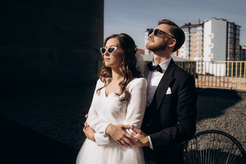 Stylishly dressed handsome groom with a beard in sunglasses hugs the beautiful bride. just married. Stylish european couple on their wedding day. wedding portrait of a couple