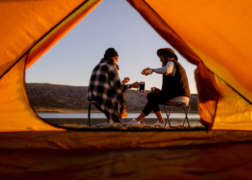 two young girlfriends traveler took a break, sit in front of the lake and drink hot drinks near the tent
