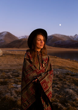 interesting girl traveler in a hat and a patterned poncho in the middle of wildlife, snowy mountains, lake, moon, beautiful sunset light
