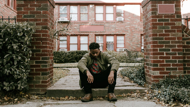 Man in green jacket and brown boots standing outside and sitting on stoop