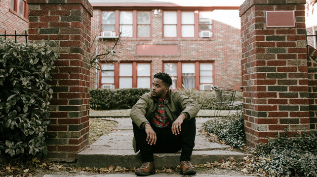 Man in green jacket and brown boots standing outside and sitting on stoop