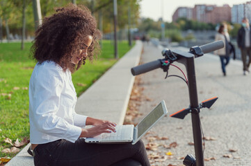 young woman sitting on a bench with a laptop and an electric scooter © Rufino